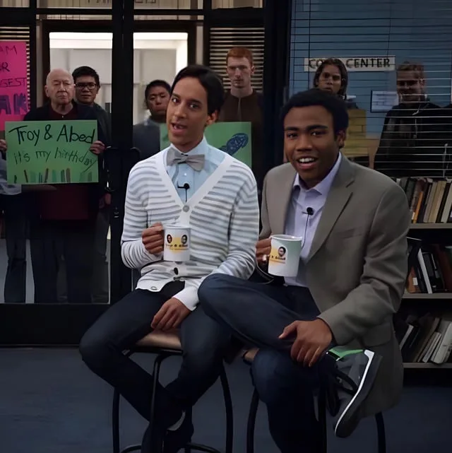 Two men sit on stools in what appears to be a college common area, holding mugs with their faces on them. Behind them, a…