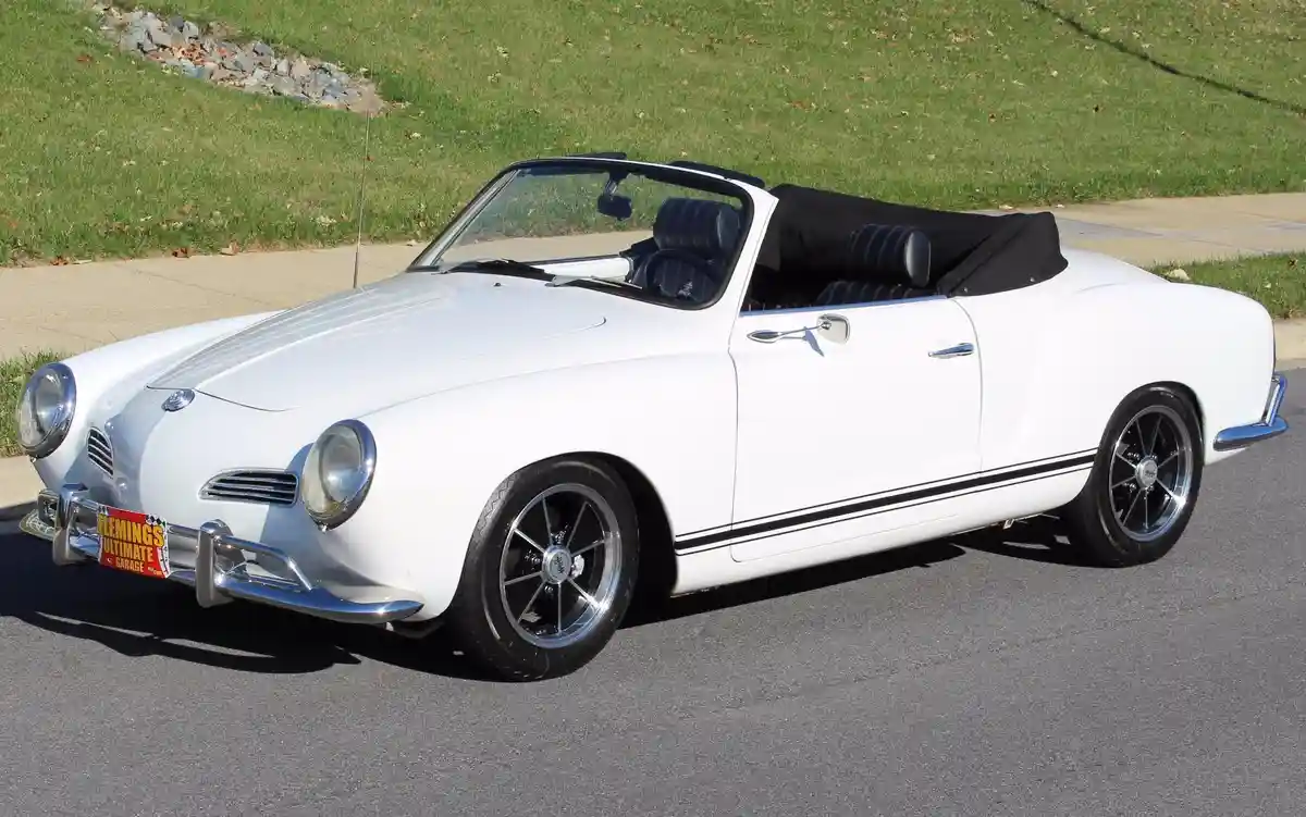 A white vintage Volkswagen Karmann Ghia convertible with black interior and side stripes is parked on asphalt, facing left…