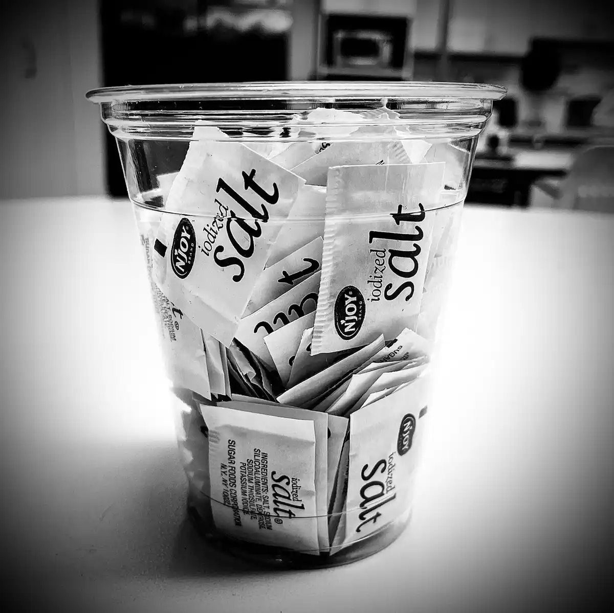 A black-and-white photo shows a clear plastic cup filled with numerous small, individual packets labeled 'iodized salt' from…