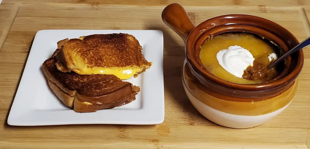 A grilled cheese sandwich cut in half and served on a white plate sits next to a brown ceramic bowl of soup topped with a…