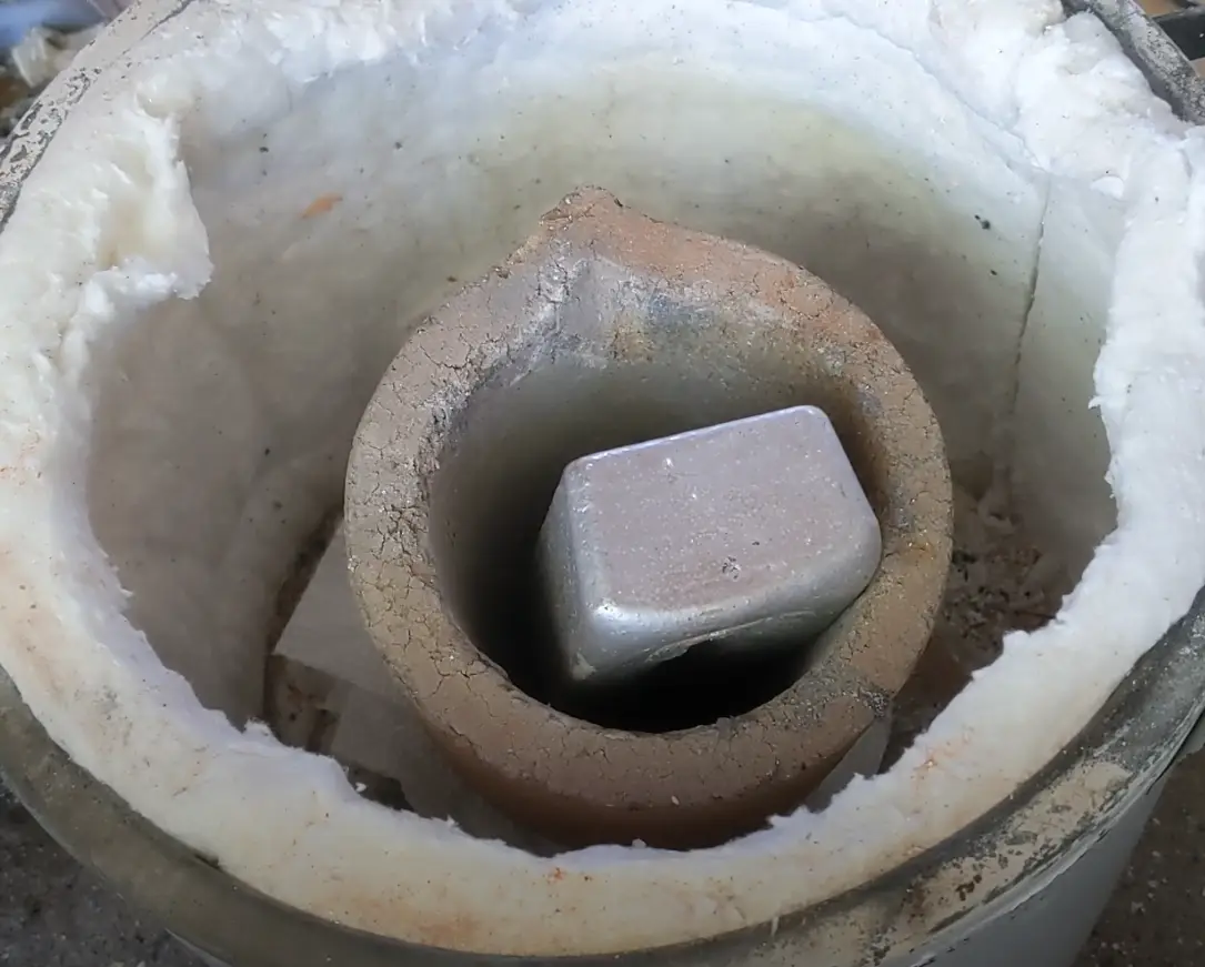A close-up photo shows a shiny, silver-colored metal ingot sitting inside a small, cracked ceramic crucible, which is placed…