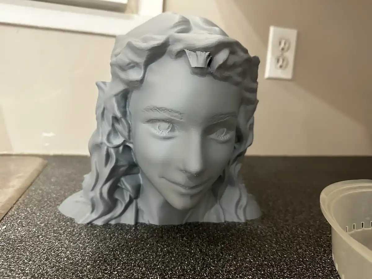 A gray 3d-printed bust of a person's head with wavy hair sits on a speckled countertop, showing a small, jagged print flaw…