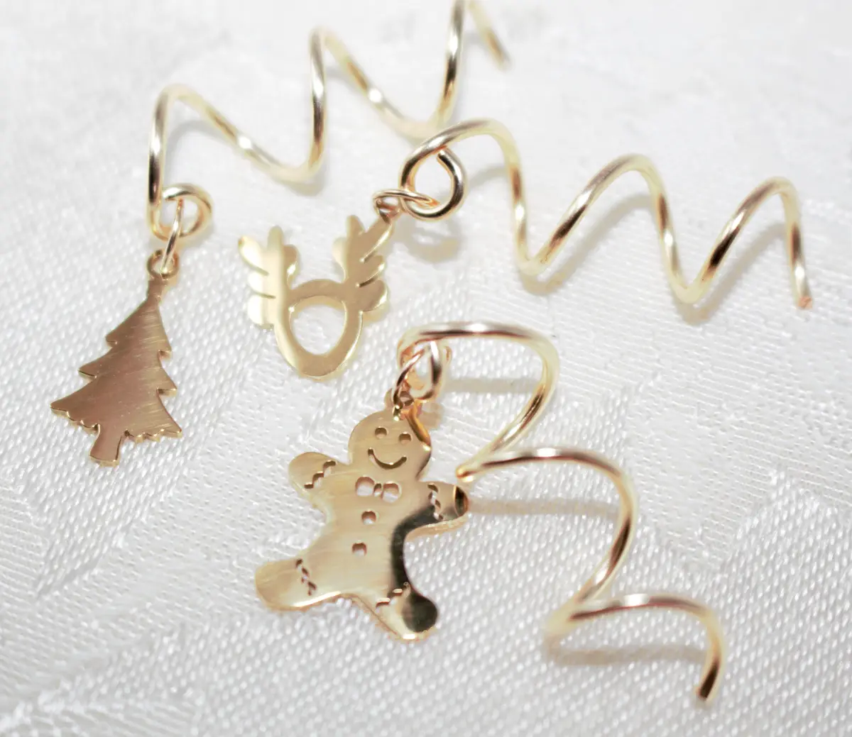 Three gold-colored Christmas-themed charms—a pine tree, a reindeer, and a smiling gingerbread man—are attached to spiral…
