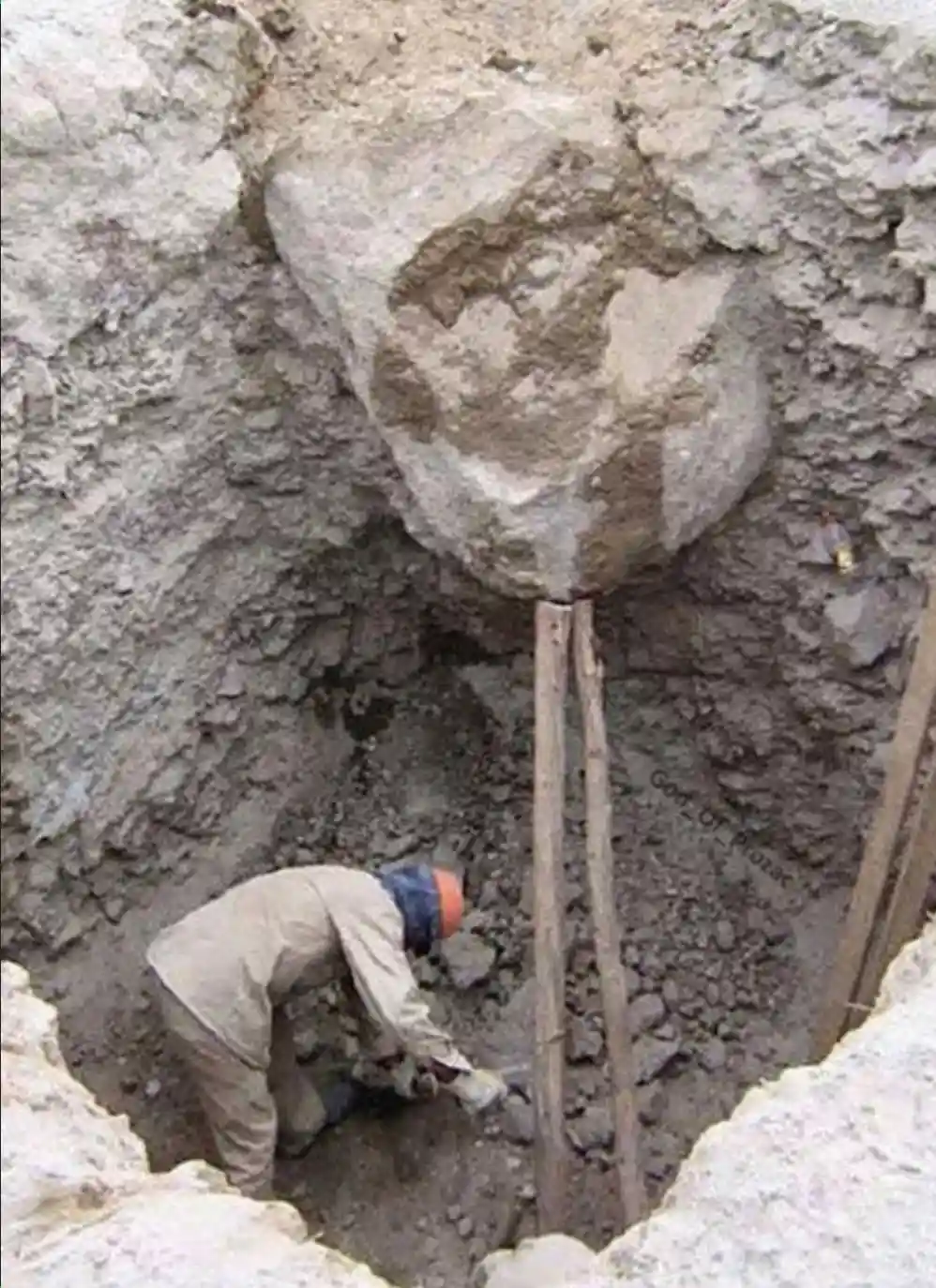 A person in a work uniform and orange hard hat is bent over digging in a deep hole, with a large, oddly shaped rock…