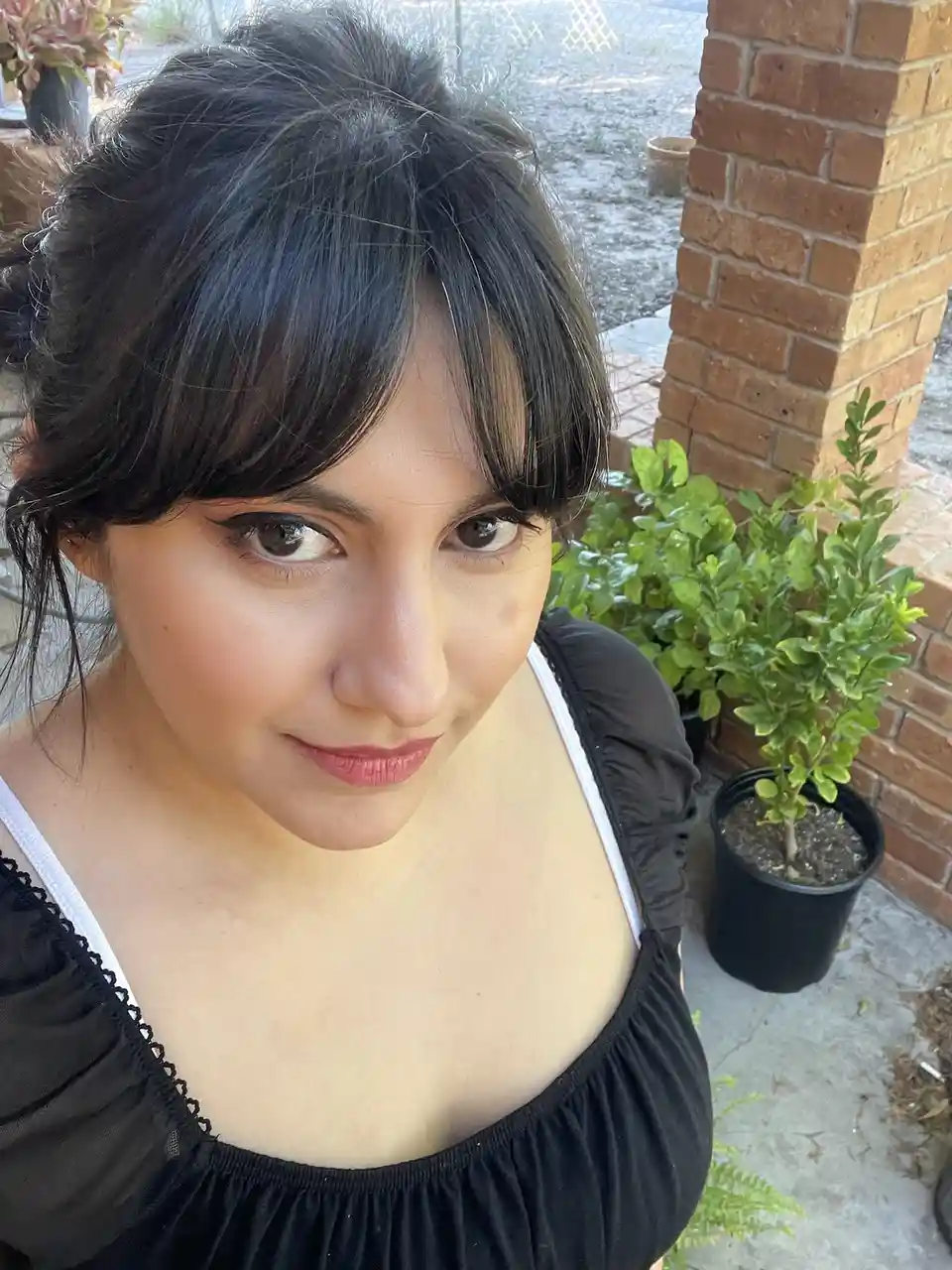 A woman with dark hair and bangs, wearing a black top with white straps, looks directly at the camera with a slight smirk…