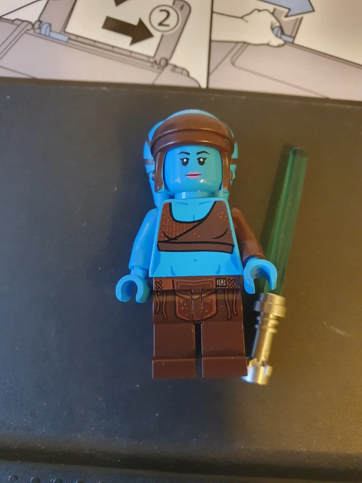 A blue-skinned LEGO minifigure resembling Rey from Star Wars, wearing brown headband and outfit, holding a green lightsaber…
