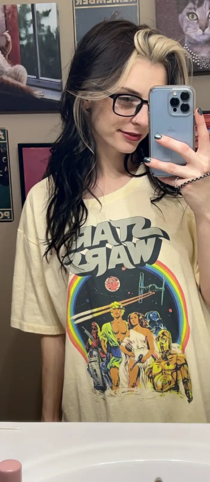 A person with dark hair and blonde highlights, wearing glasses and a beige t-shirt with a retro 'Star Wars' graphic…