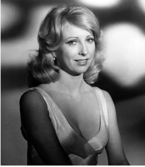 A black-and-white portrait of a woman with blonde hair and a slight smile, wearing a low-cut dress. The text overlaid on the…