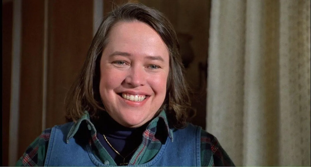 A woman with shoulder-length brown hair, wearing a plaid shirt under denim overalls and a dark turtleneck, smiles broadly at…