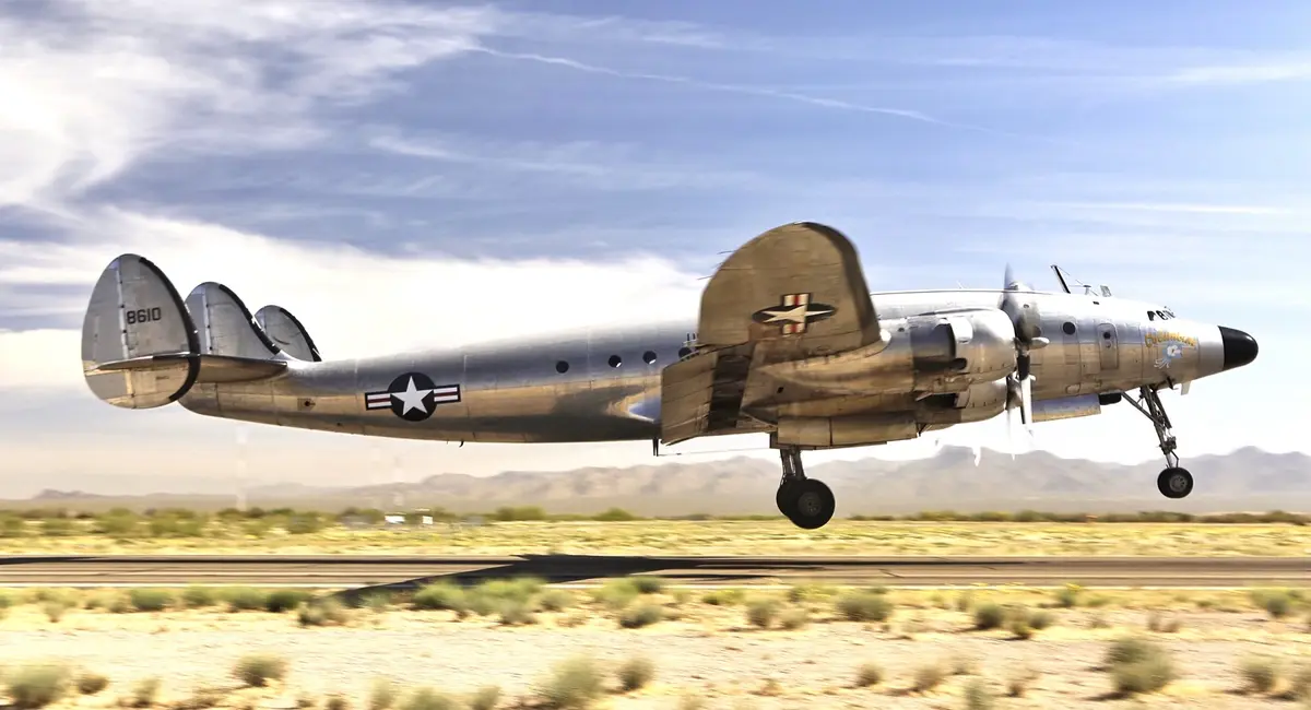 A silver WWII-era military transport plane, likely a C-47 Skytrain, is captured mid-takeoff over a desert runway with…