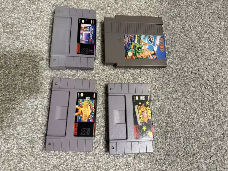 Four Super Nintendo cartridges — EarthBound, Kirby Super Star, Island, and a game with a black label — lie on a gray carpet…