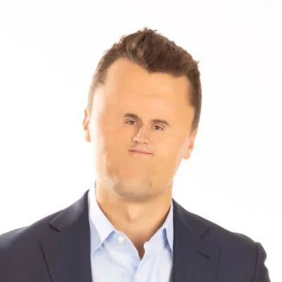 A man in a dark suit and light blue shirt makes a skeptical, slightly disgusted facial expression while looking directly at…