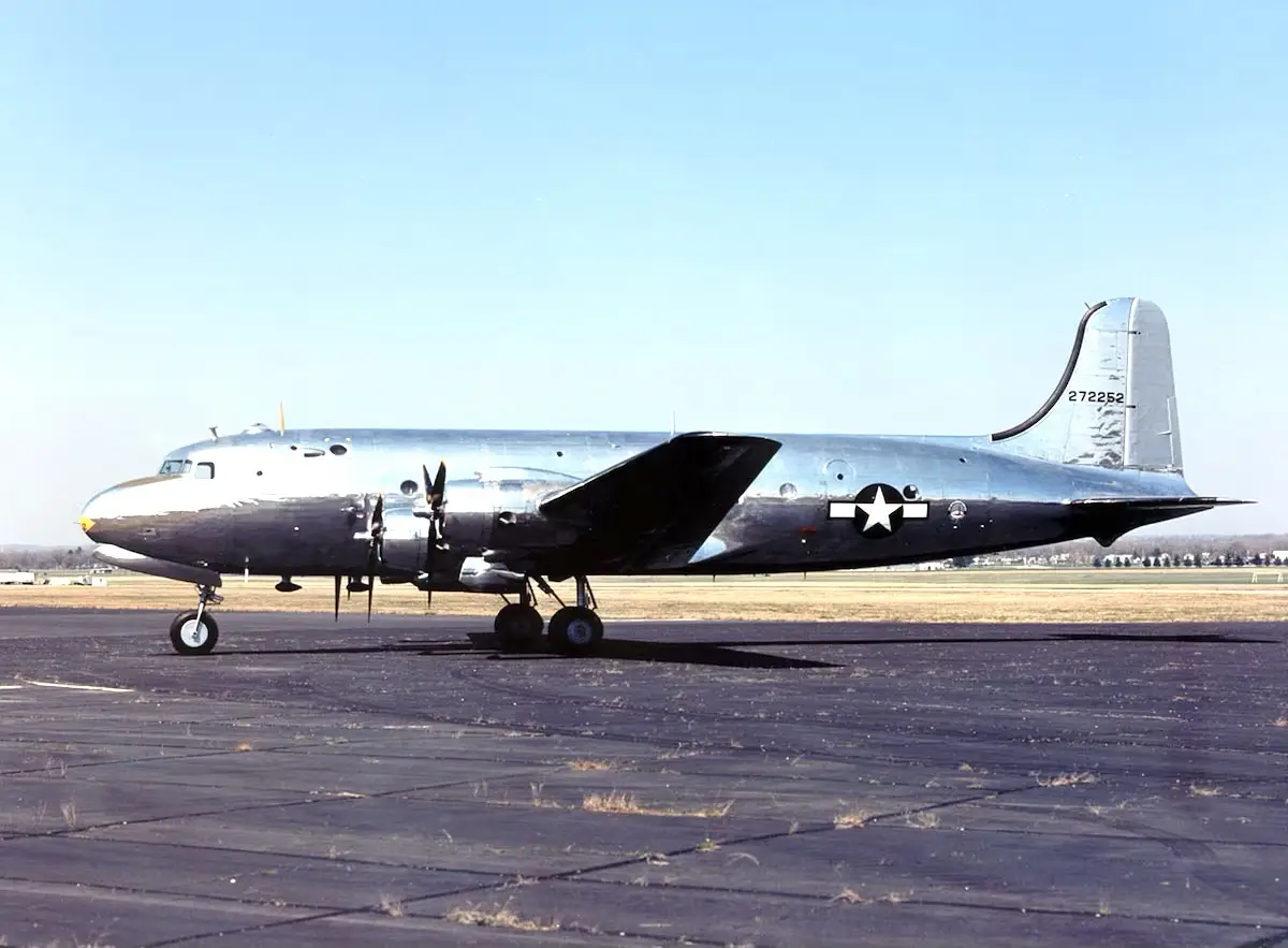 A silver military transport aircraft, likely a C-47 Skytrain, is parked on an asphalt runway under a clear blue sky. The…