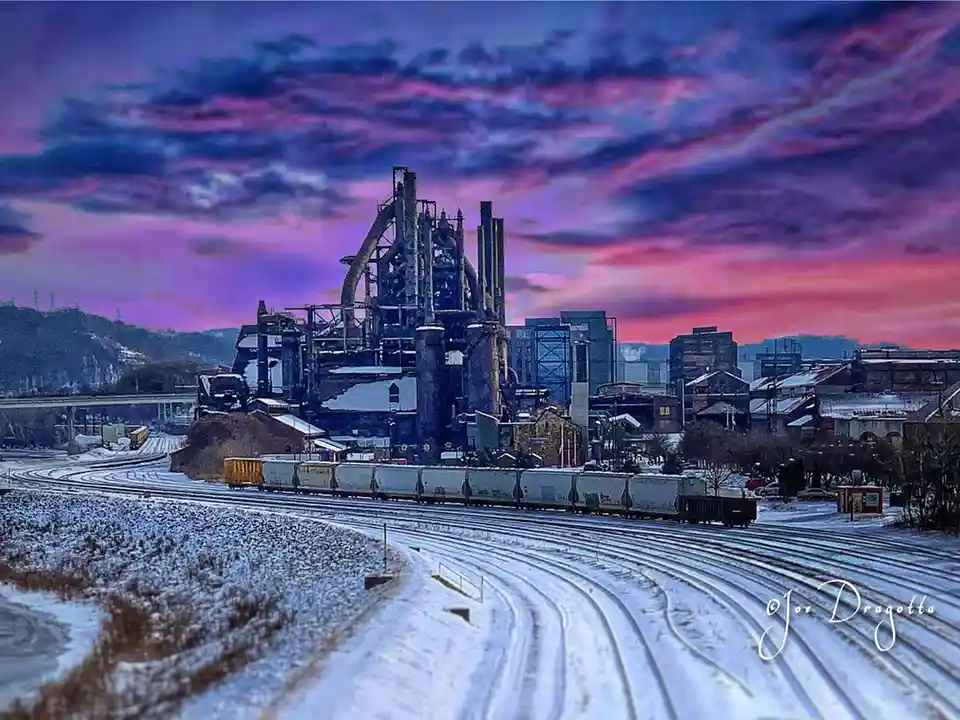 A snow-covered industrial landscape at dusk, featuring a large steel mill with intricate metal structures, multiple train…