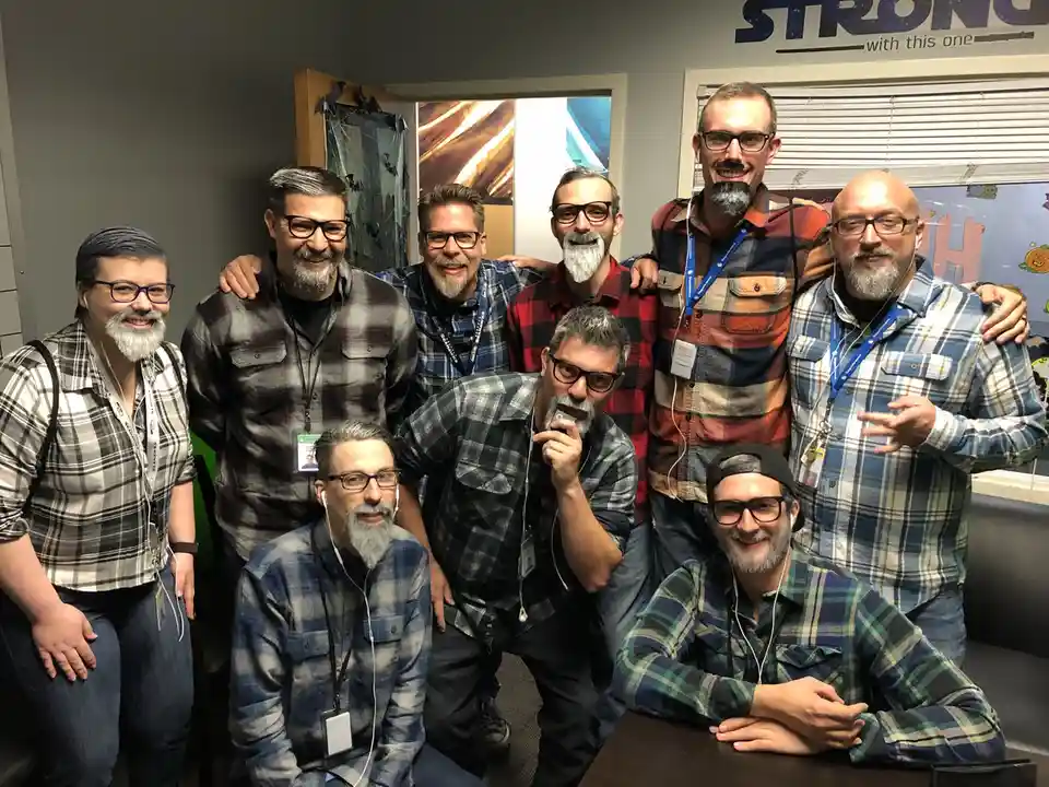 A group of eight men, mostly wearing plaid shirts and glasses, pose together in an office setting. All of them have…