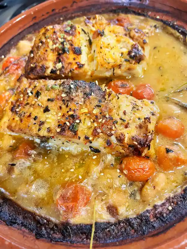 A close-up photo of a cooked fish dish in a terracotta tagine pot, featuring golden-brown fish fillets, small orange…