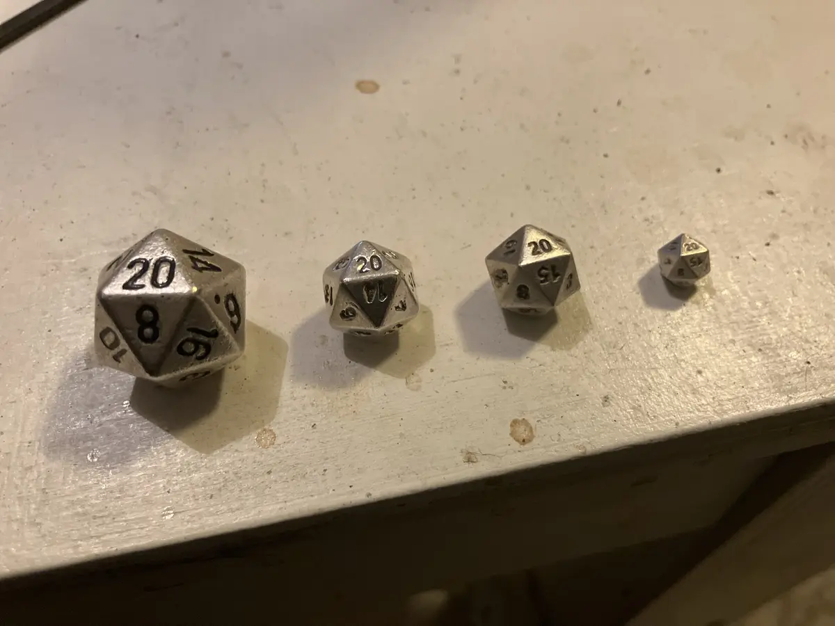 Four metallic 20-sided dice are arranged in a row on a white, slightly worn surface, decreasing in size from left to right…