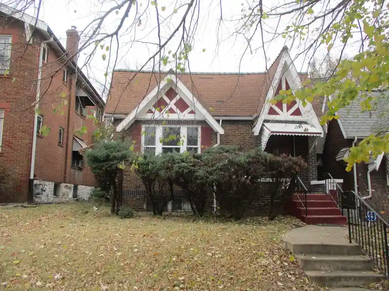 A photo of a brick house with red and white decorative gables, surrounded by bushes and fallen leaves in autumn. The…