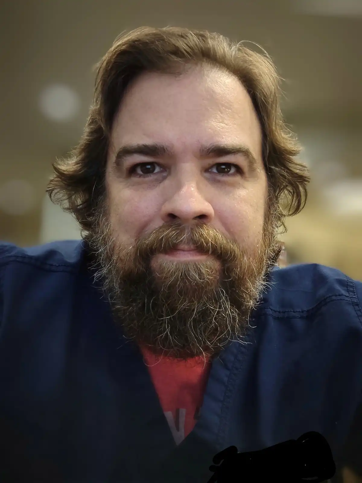 A close-up photo of a man with a full brown beard and mustache, wearing a blue jacket over a red shirt, looking directly at…
