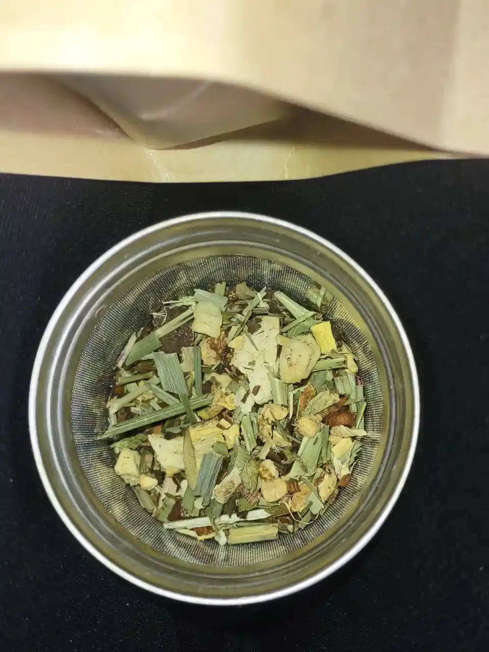 A top-down view of a metal tea strainer filled with dried green tea leaves and other botanicals, resting on a black surface…