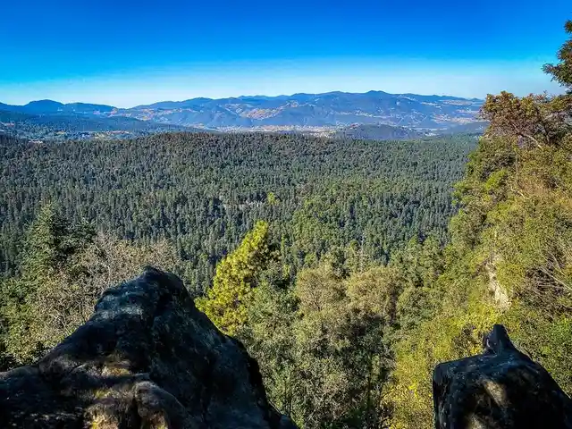 A scenic photograph shows a vast forested valley with distant mountains under a clear blue sky, framed by large dark rocks…