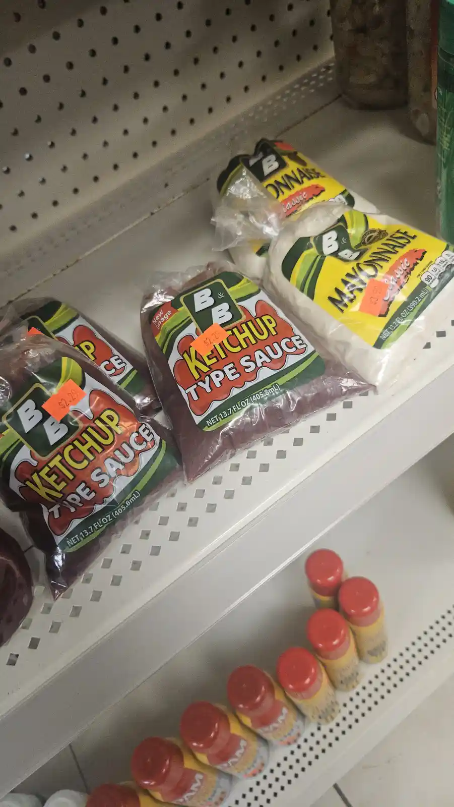 A grocery store shelf displays bags of 'B&B KETCHUP TYPE SAUCE' and 'B&B MAYONNAISE' with orange price tags, humorously…