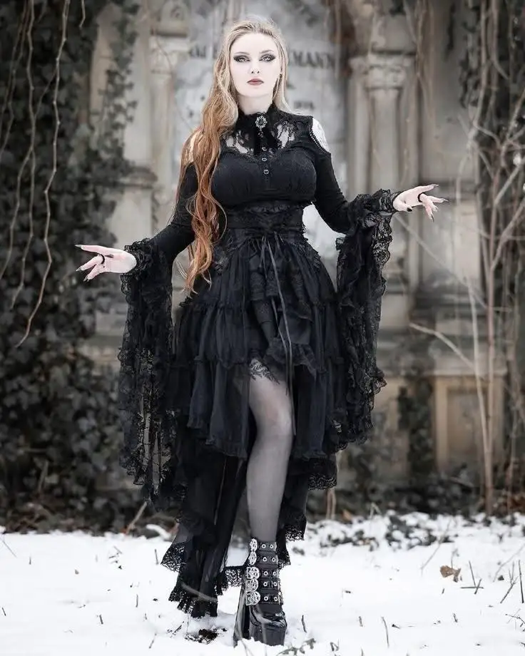 A woman with long blonde hair stands in a snowy graveyard wearing an elaborate black gothic dress with lace, ruffles, and a…