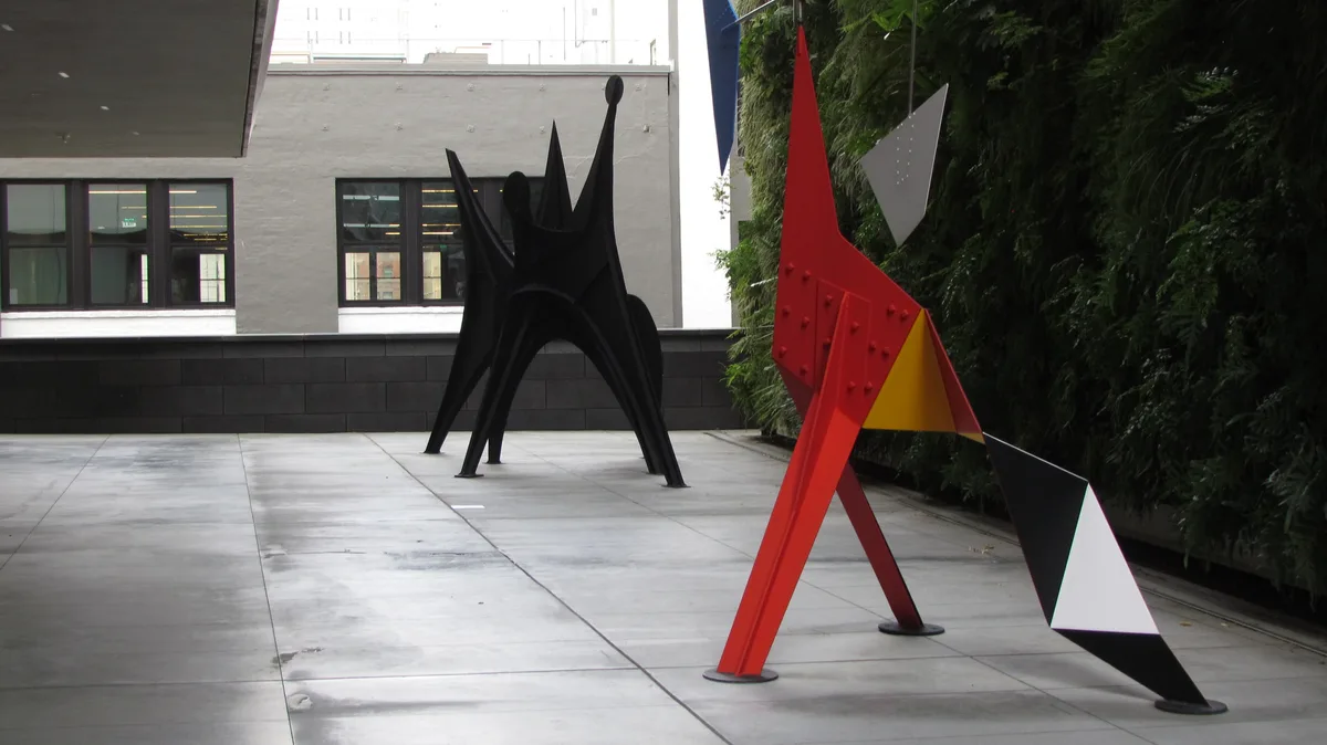 Two abstract sculptures stand in an outdoor courtyard: a black, spiky, humanoid figure and a red, yellow, black, and white…