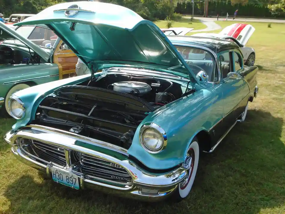 A vintage turquoise and black Oldsmobile with its hood open, parked on grass at a car show with other classic cars nearby…