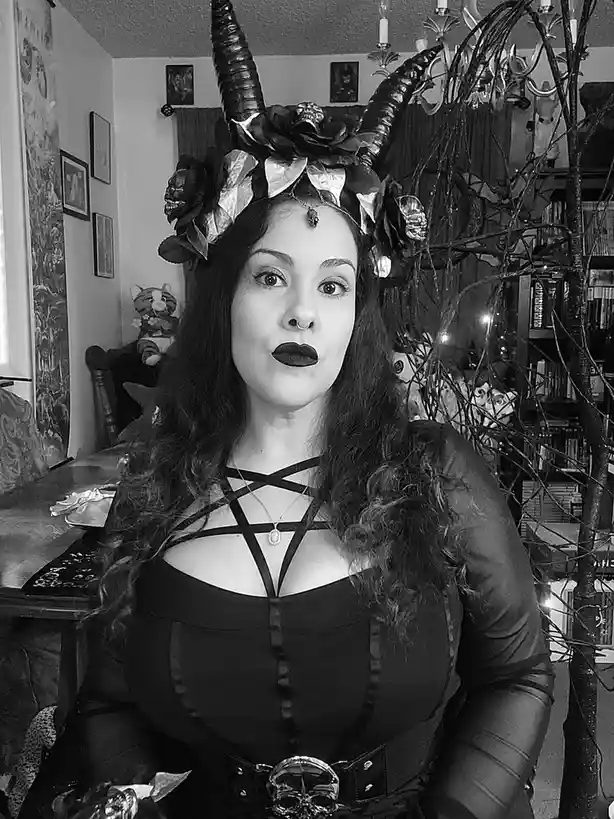 A black-and-white photo of a woman dressed in a gothic devil costume with large horns, dark makeup, and a corset, looking…