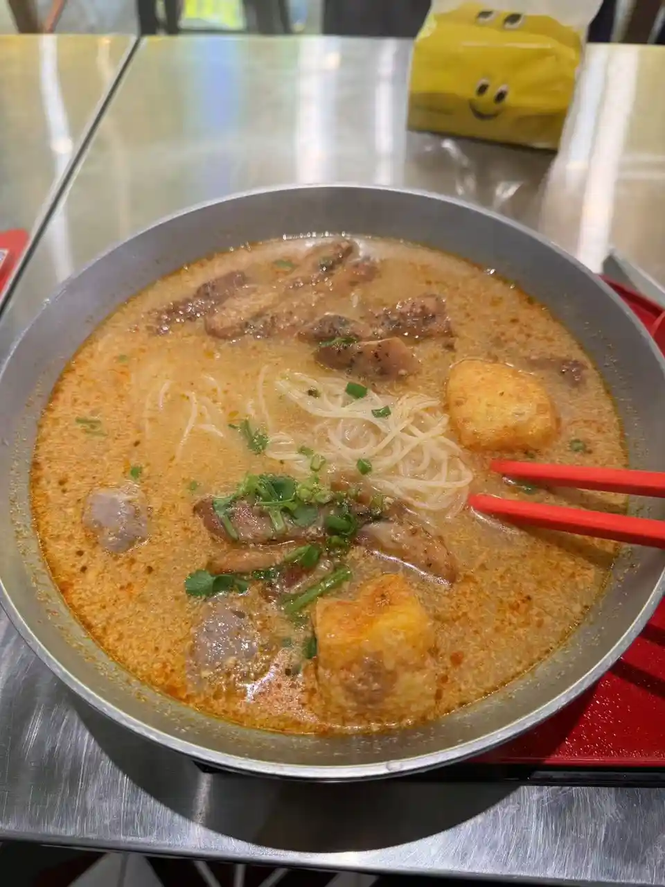 a close-up photo shows a metal bowl filled with a creamy, orange-colored ramen broth containing noodles, meat slices, and…
