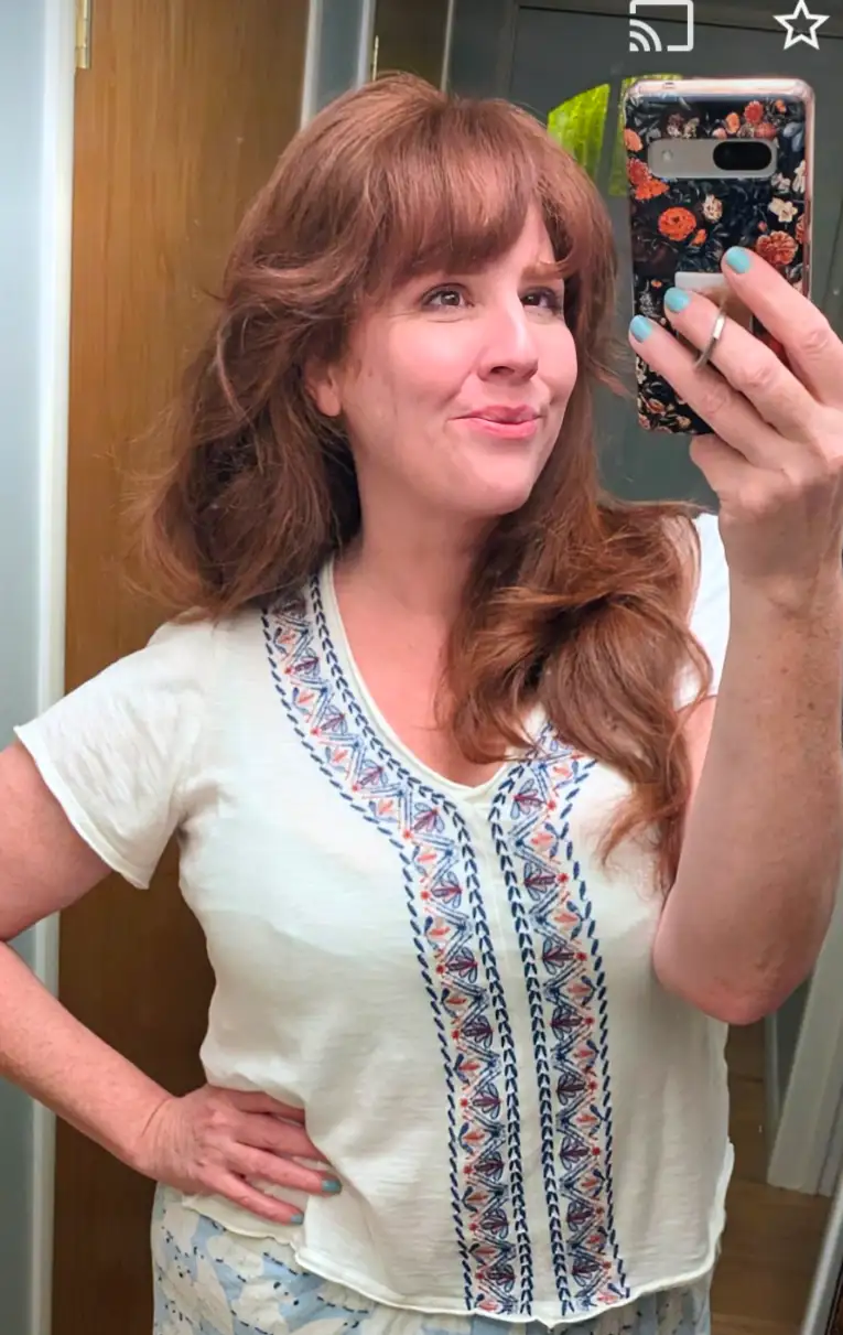A woman with reddish-brown hair and light blue nail polish takes a mirror selfie while wearing a white short-sleeved shirt…
