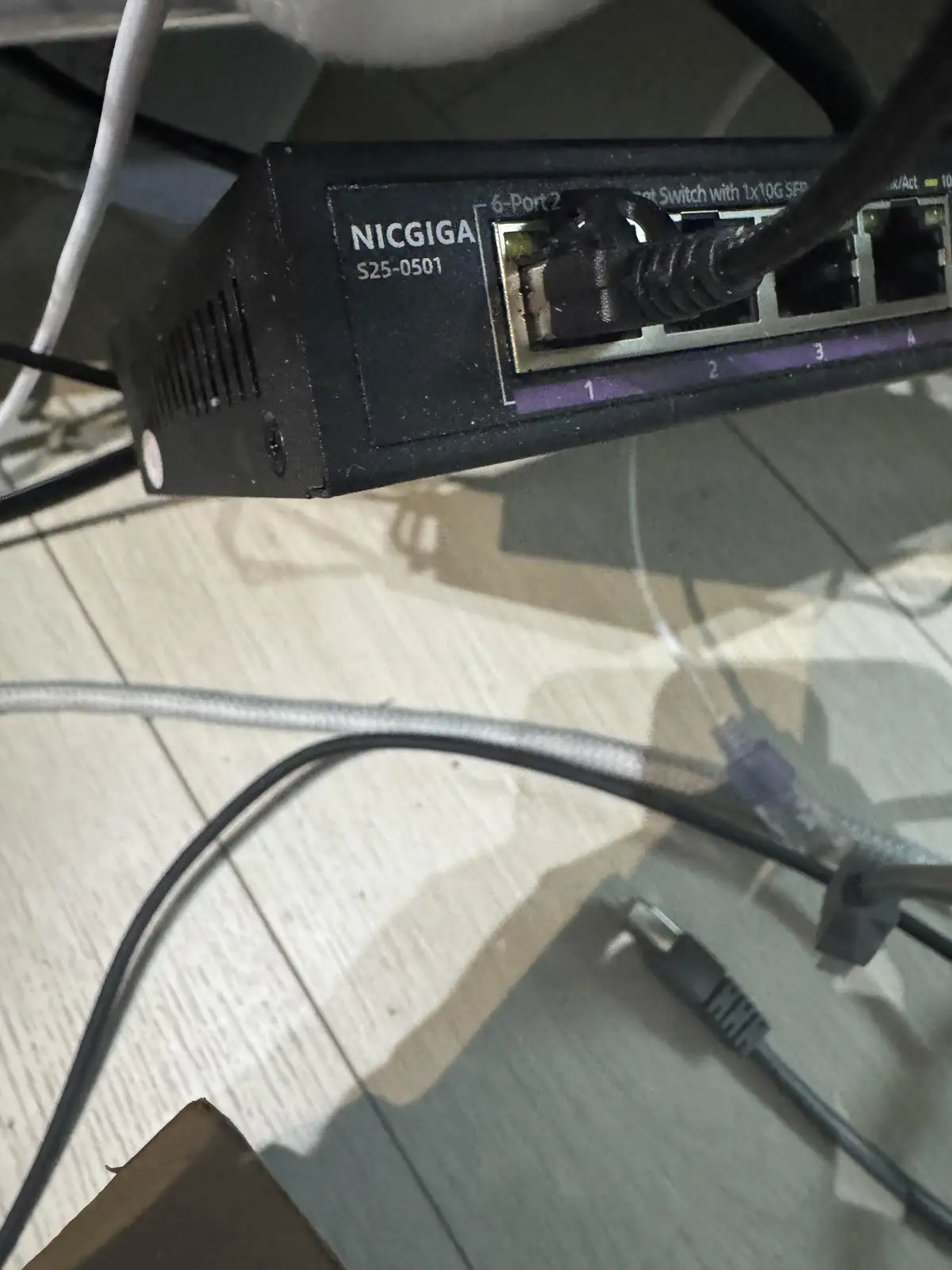 A close-up photo shows a black NICGIGA network switch with a cable plugged into port 1, sitting on a tiled floor with other…