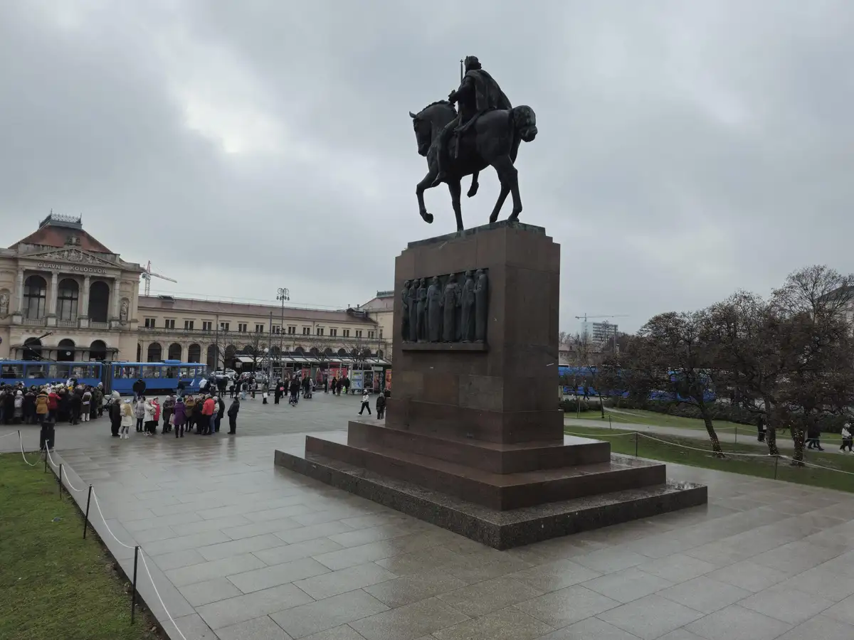 A large equestrian statue of a historical figure stands on a stone pedestal in a public square, with people and blue trams…