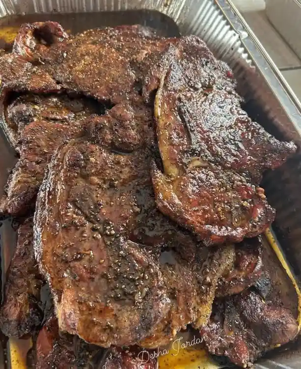 A close-up photo shows a large metal tray filled with numerous thick, juicy, well-seasoned grilled steaks, glistening with…