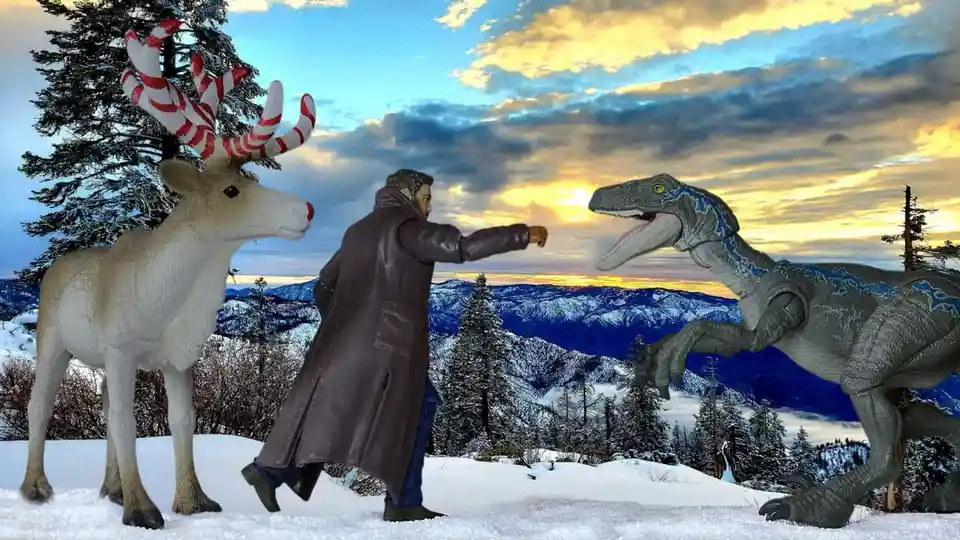 A toy figure of a man in a brown coat stands in a snowy mountain landscape, pointing at a toy velociraptor. To his left is a…