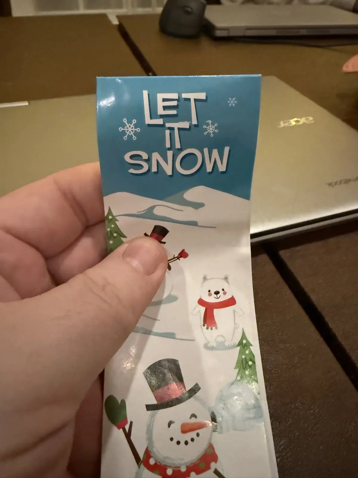 A hand holds a festive holiday bookmark with a blue top section that says 'LET IT SNOW' surrounded by snowflakes, and a…