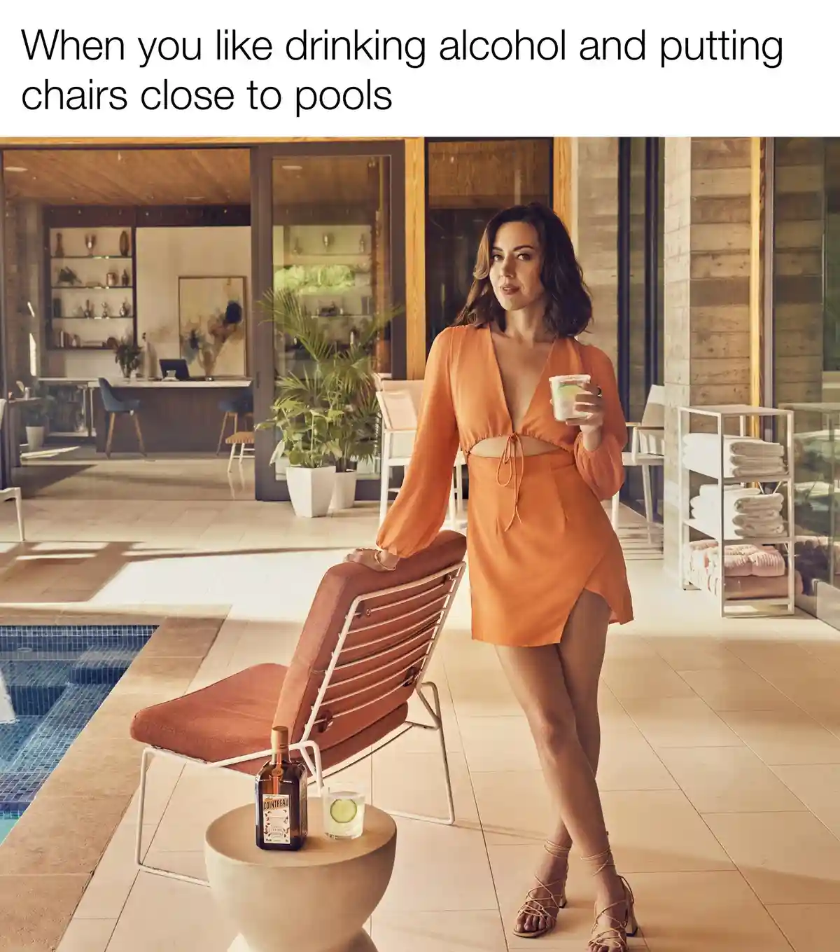 A woman in an orange dress stands beside a lounge chair placed directly next to a swimming pool, holding a cocktail. A…