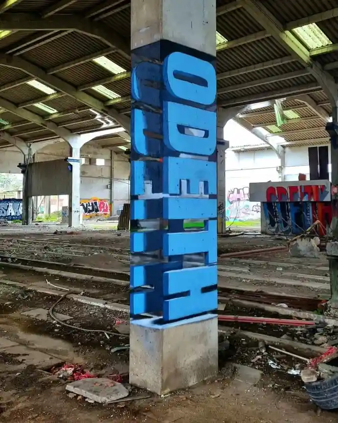 A large concrete pillar in a derelict, abandoned train station is covered in bold blue graffiti spelling 'ODE TO THE'. The…