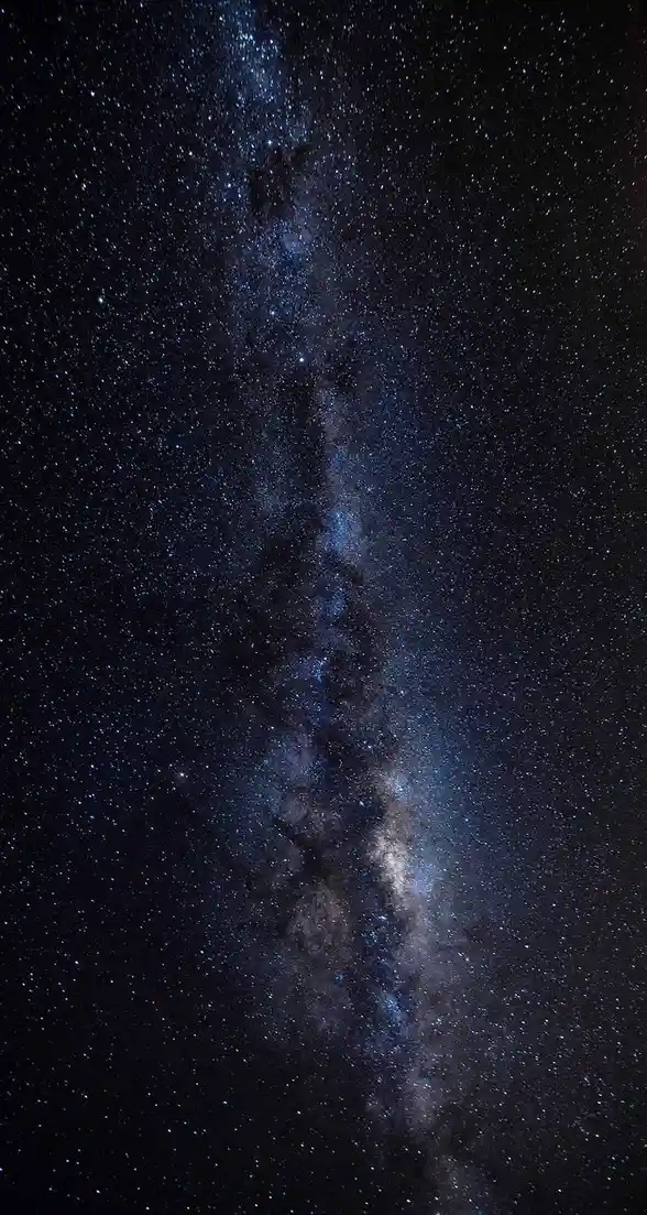 A vertical photograph of the Milky Way galaxy, showing a dense band of stars, cosmic dust, and nebulae stretching across a…