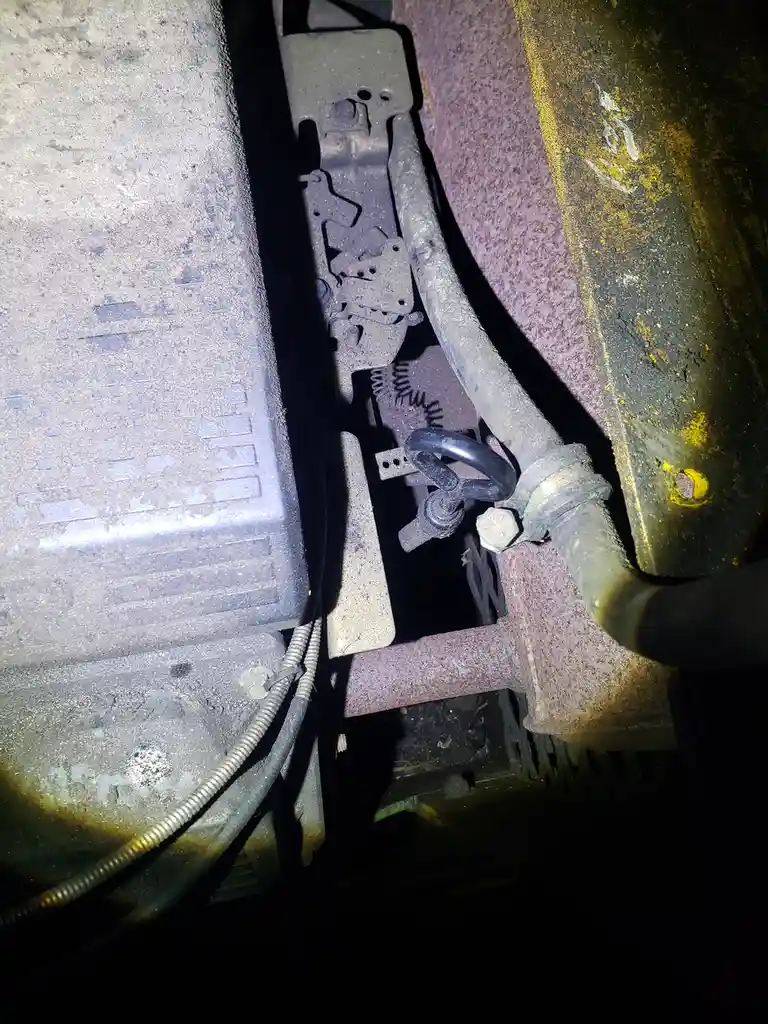 A close-up photo of a rusty, dirty undercarriage of a vehicle, showing metal brackets, wires, and a bolted pipe. A small…