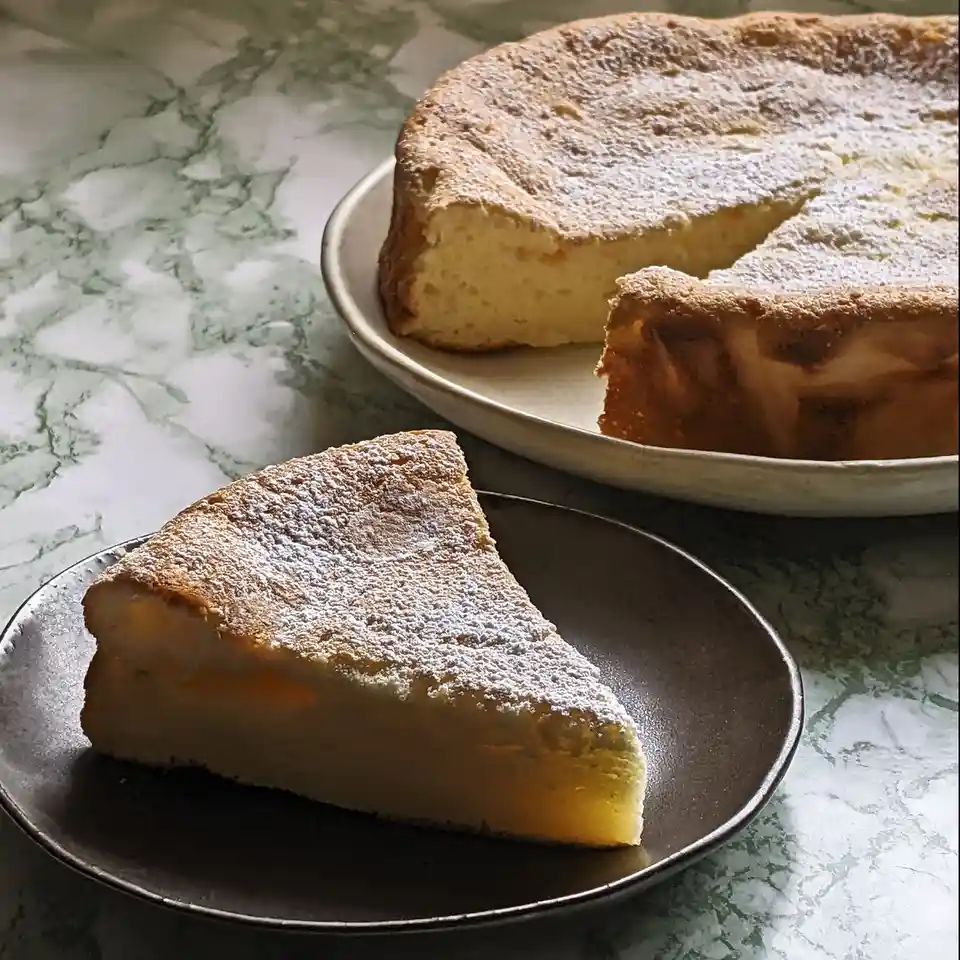 A slice of golden-brown, powdered sugar-dusted cake sits on a dark plate in the foreground, with the larger cake on a white…