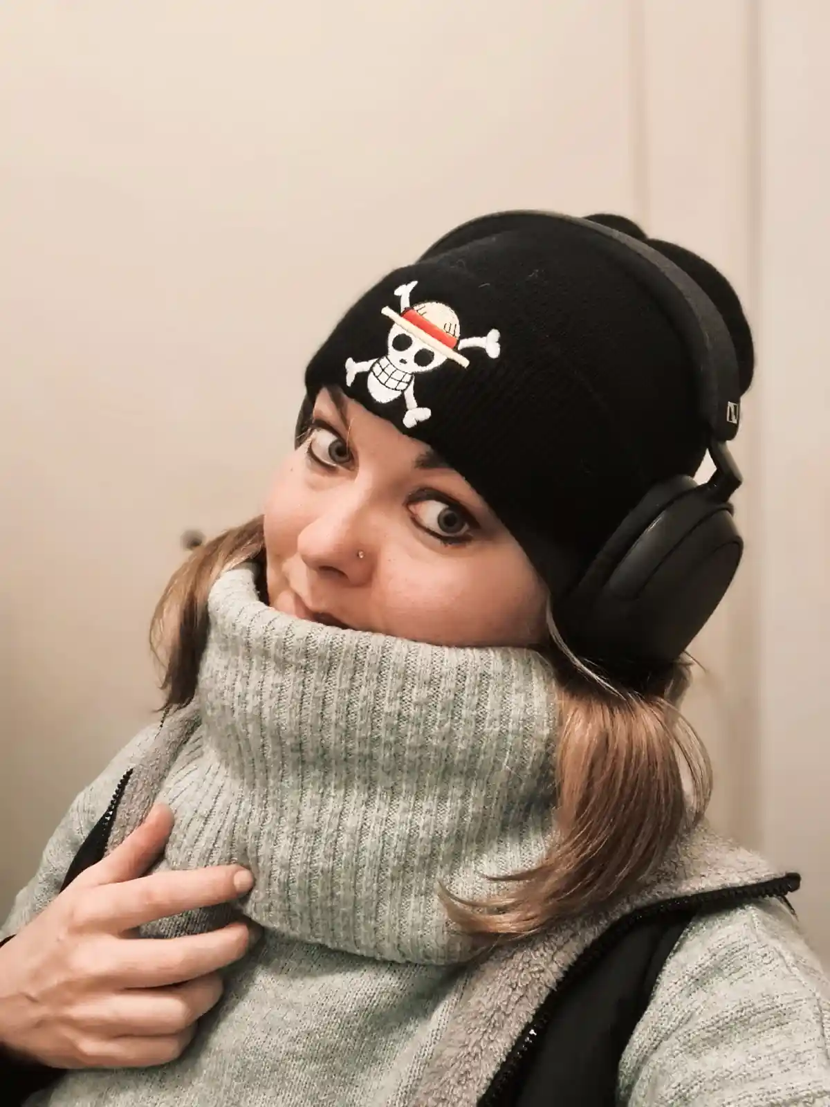 A person wearing a black beanie with a white skull and crossbones logo, large black over-ear headphones, and a high-necked…