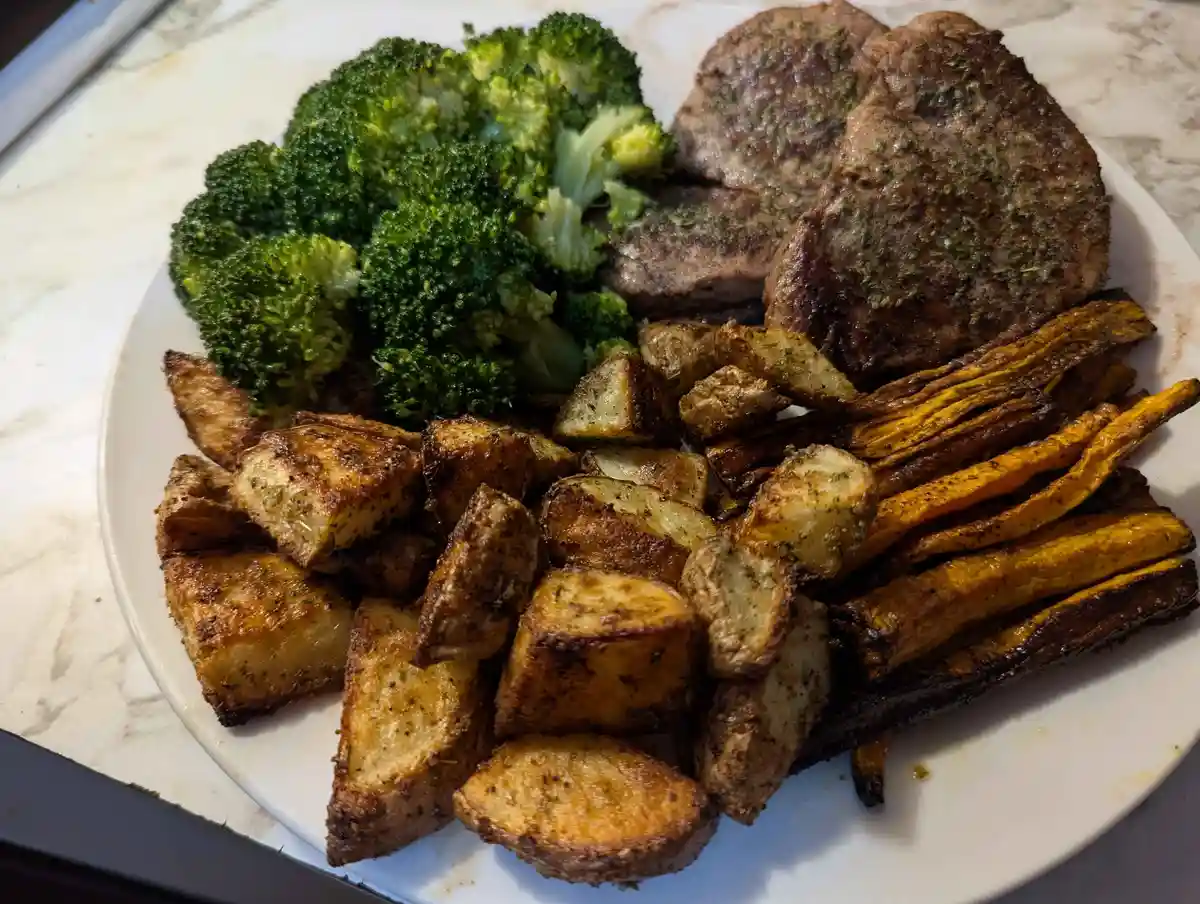 A white plate holds a meal of roasted meat, steamed broccoli, roasted potato wedges, and roasted sweet potato fries, all…