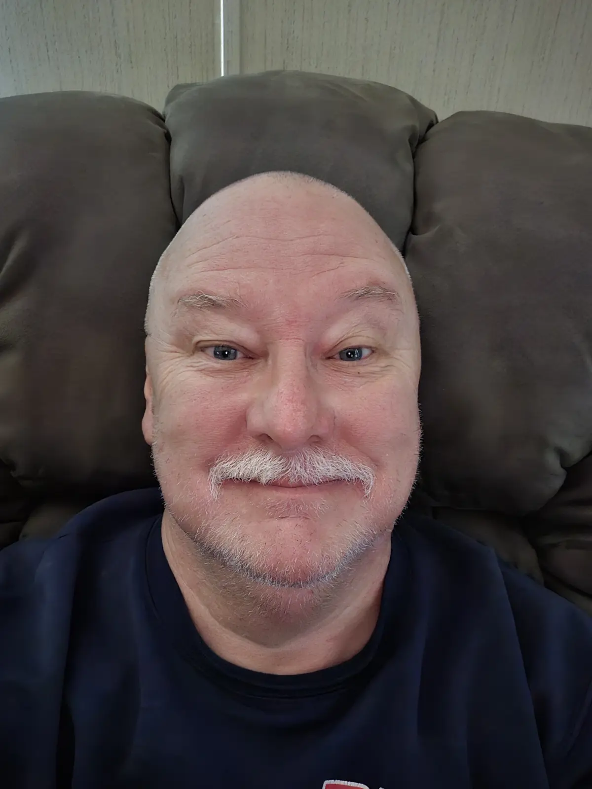 A bald man with a white mustache and goatee is lying back on a dark brown couch, smiling slightly at the camera. He wears a…