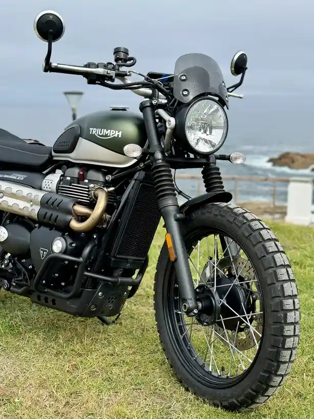 A close-up photo of a matte green Triumph Street Scrambler motorcycle parked on grass near the ocean, featuring a round…