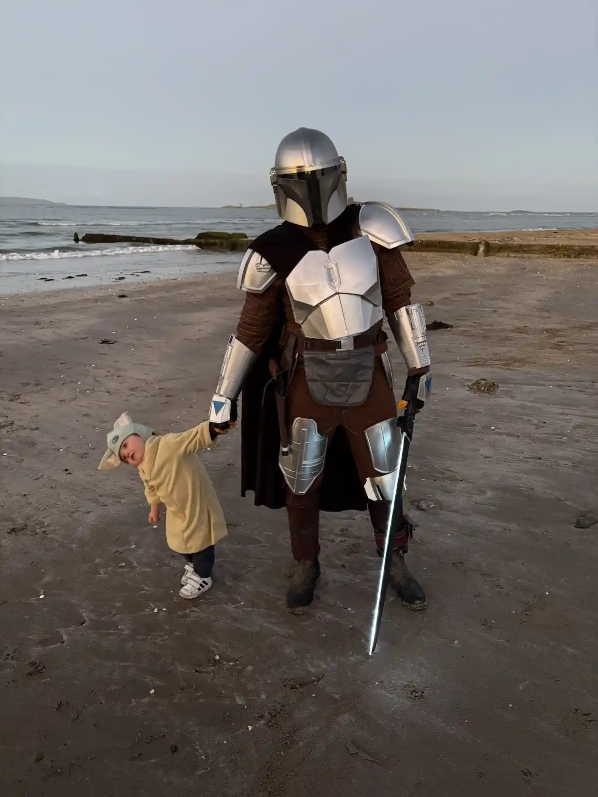 An adult in a full Mandalorian armor costume, holding a glowing lightsaber, stands on a beach holding the hand of a toddler…
