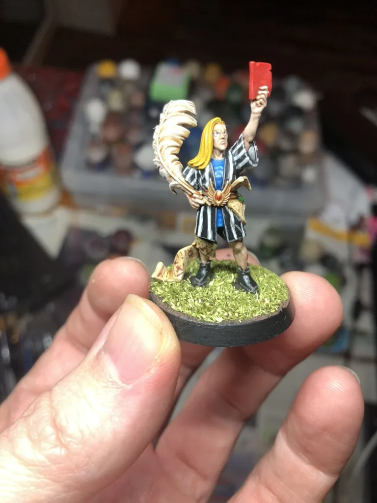 A hand holds a painted miniature figure of a fantasy character with long blonde hair, wearing a striped robe and holding a…