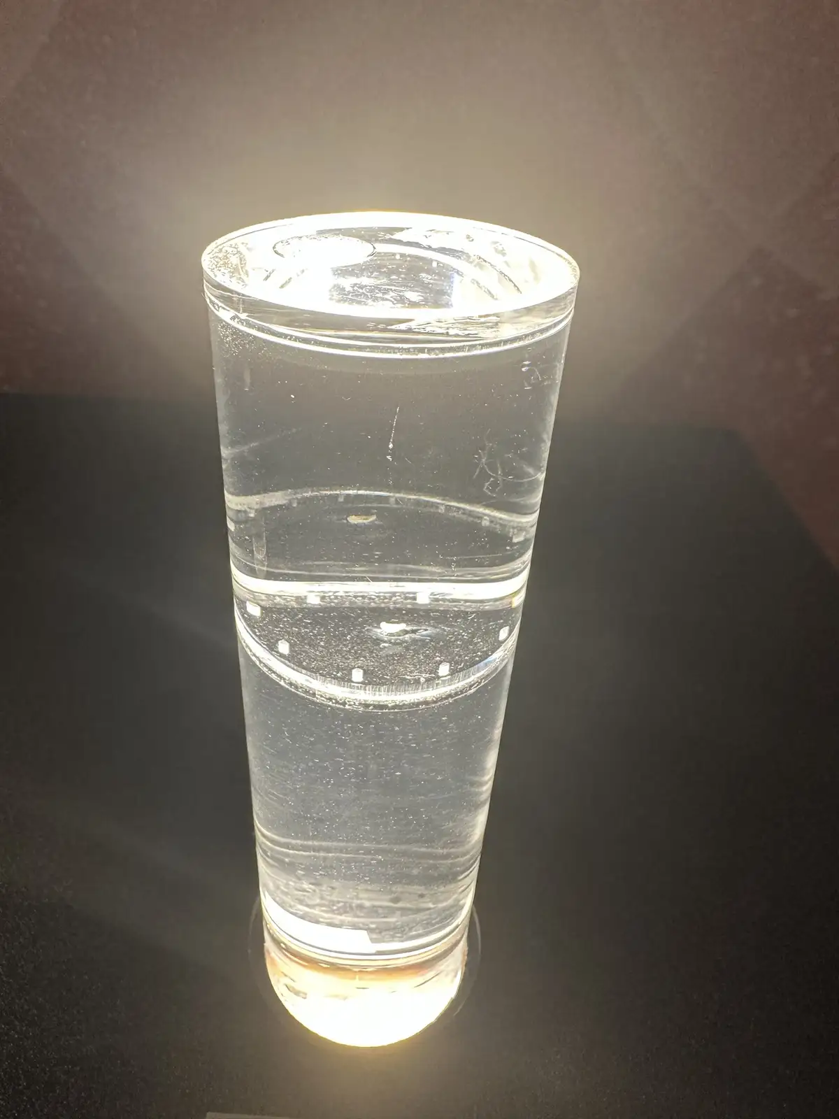 A tall, clear glass filled with water sits on a dark surface, illuminated from below and above, creating bright highlights…