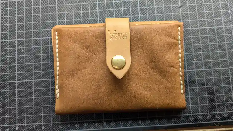 A handmade tan leather wallet with white stitching and a brass snap closure rests on a gray cutting mat with grid lines and…